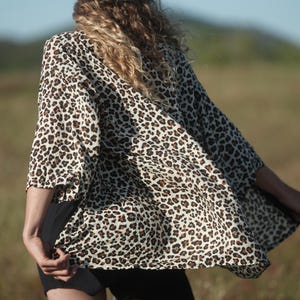 May include: A woman wearing a black hat and a leopard print kimono-style top. The top is cream-colored with brown spots. The woman is standing in a field of tall grass.