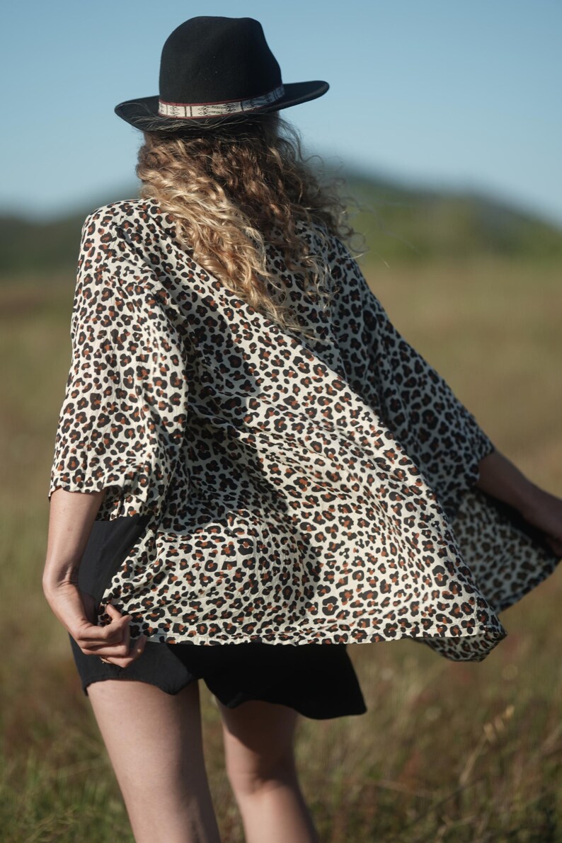 May include: A woman wearing a black hat and a leopard print kimono-style top. The top is cream-colored with brown spots. The woman is standing in a field of tall grass.