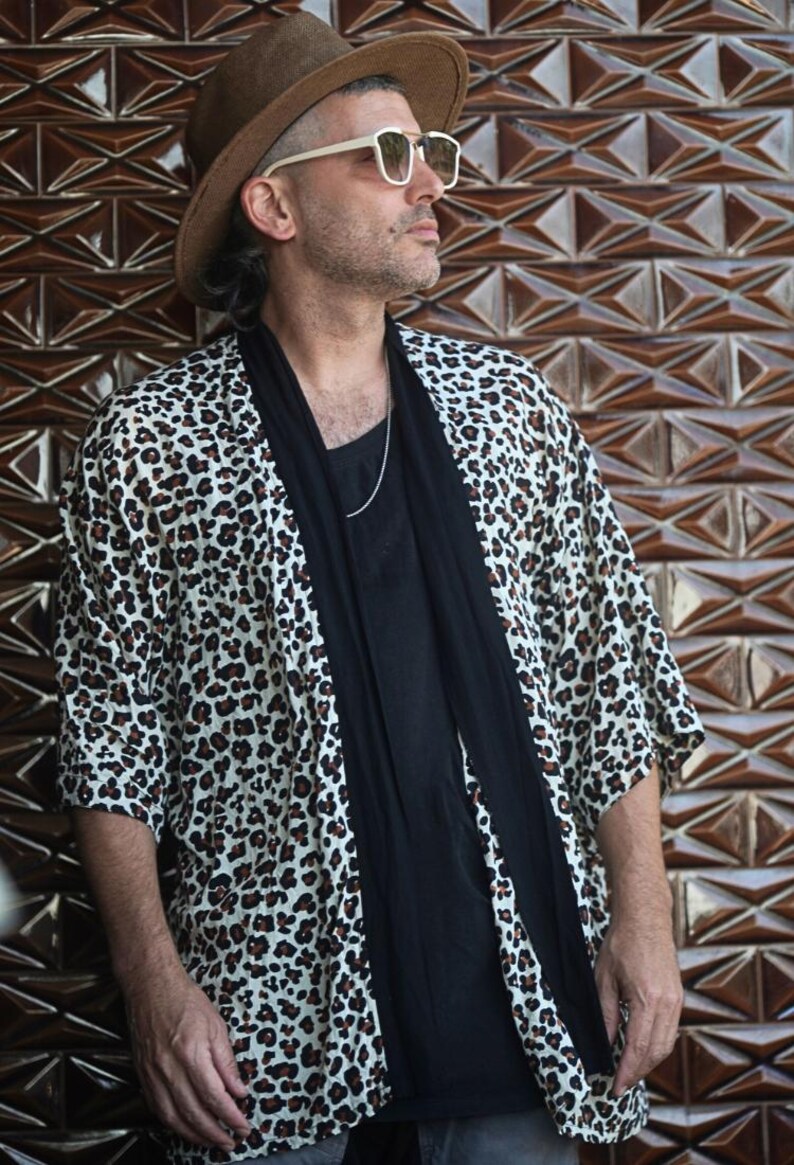 May include: A person wearing a brown hat, white-framed sunglasses, and a leopard print kimono-style jacket over a black shirt. A black scarf is draped around the neck. The background is a brown geometric patterned wall.