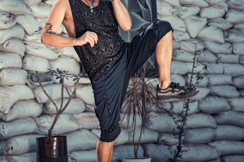 May include: A black sleeveless tank top featuring a geometric gold-toned pattern paired with black cargo-style shorts that have side pockets and dark sneakers with brown accents. The outfit is worn by a person in a dynamic pose with one leg raised.