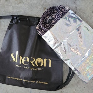 May include: A black drawstring bag with the gold text "sheron" and "WEAR IT LIKE YOU MEAN IT" alongside a website and social media handles. Next to it is a silver holographic pouch containing a shirt with a dark purple and white hexagon pattern.