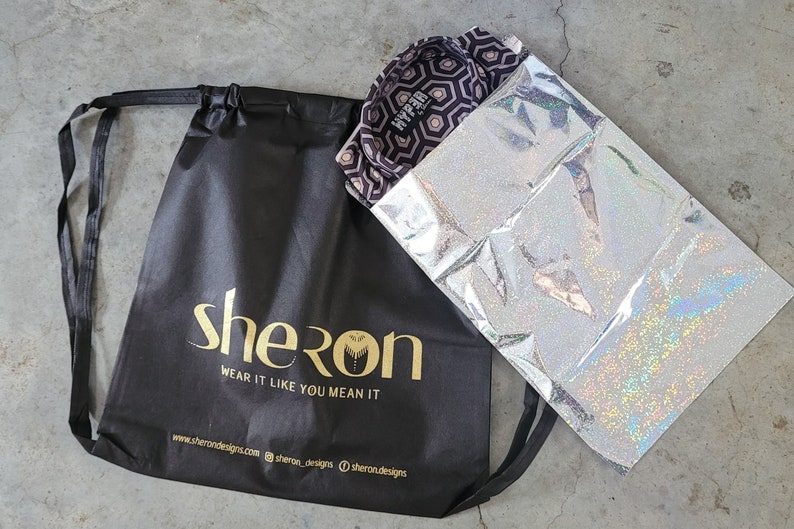 May include: A black drawstring bag with the gold text "sheron" and "WEAR IT LIKE YOU MEAN IT" alongside a website and social media handles. Next to it is a silver holographic pouch containing a shirt with a dark purple and white hexagon pattern.