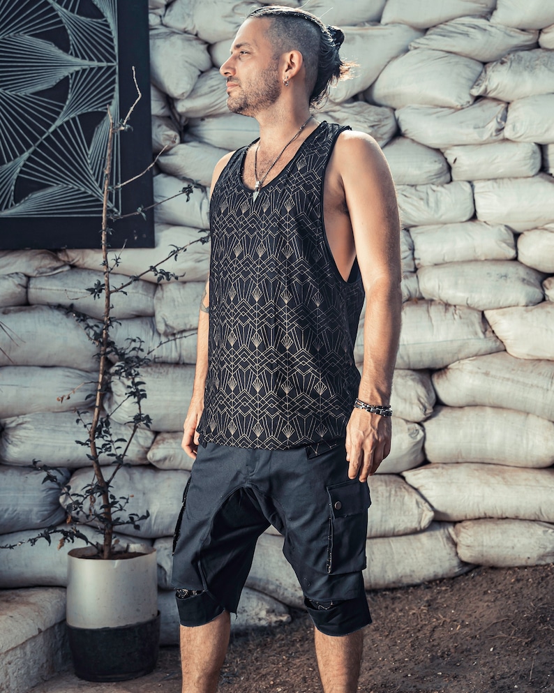May include: A man wearing a black tank top with a gold geometric pattern and black cargo shorts. He is standing in front of a wall made of white sandbags.