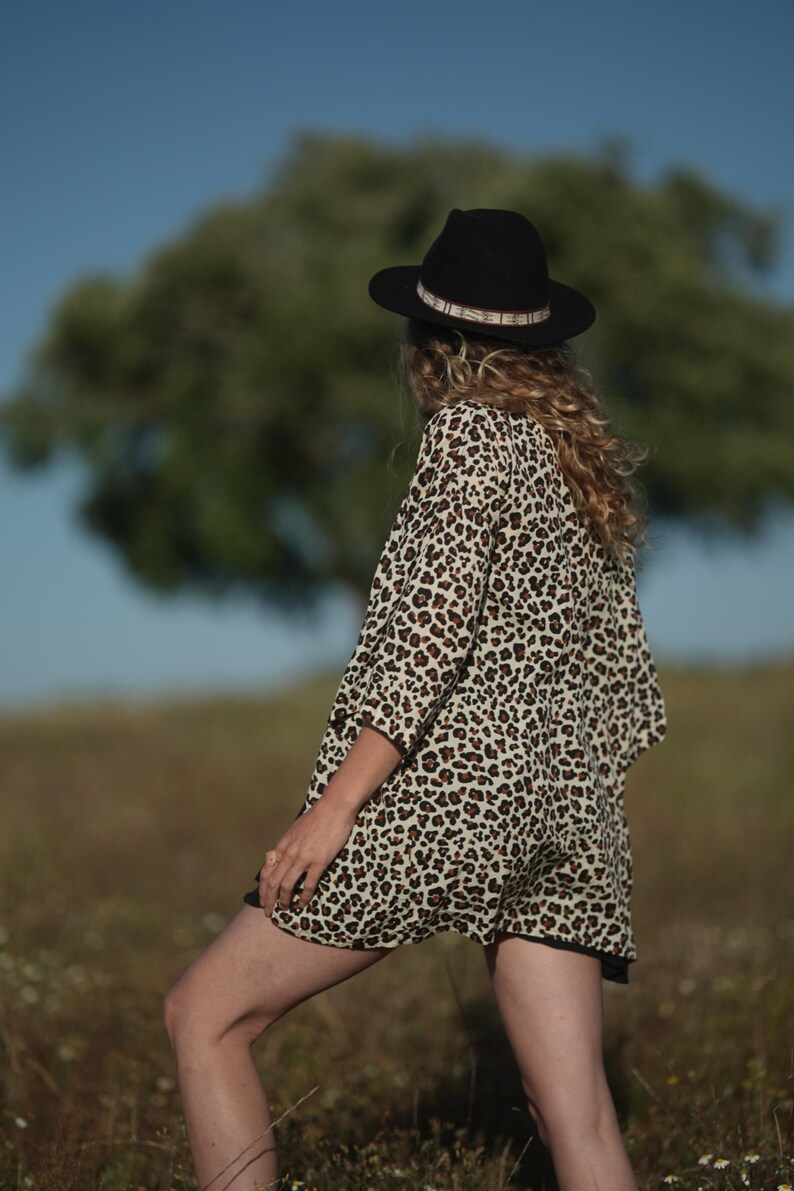 May include: A person wearing a black hat with a patterned band and a leopard print top and shorts. The outfit is in a field with a tree in the background. The outfit is a neutral color with black accents.