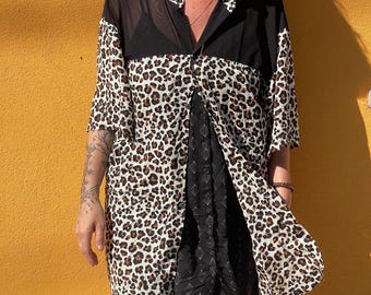 Op de afbeelding: Een button-down shirt met een luipaardprint in bruin en zwart, gecombineerd met een zwarte jurk. Het shirt heeft een doorschijnend zwart paneel aan de bovenkant en een kraag. De jurk eronder heeft een ruitpatroon.