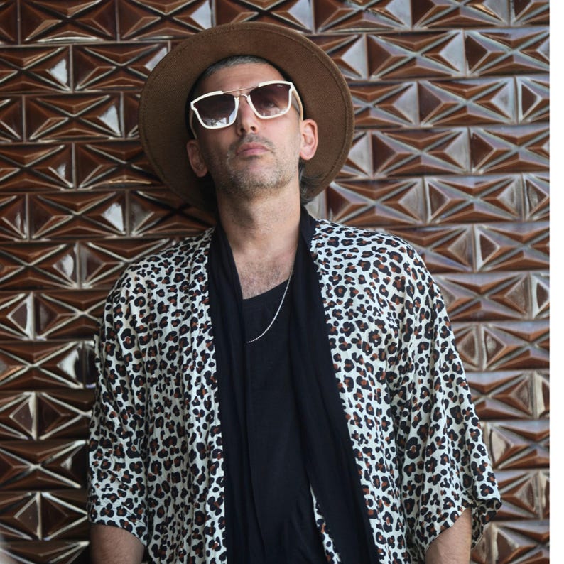 May include: A person wearing a brown hat, sunglasses, and a leopard print kimono-style jacket. The jacket is white with brown spots. The person is wearing a black shirt and a silver chain necklace.