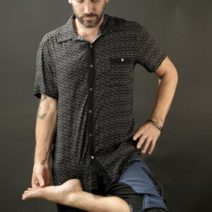 May include: A man wearing a black and white patterned short-sleeved shirt and blue trousers with a geometric design. He is standing with one leg bent and his foot resting on his thigh.