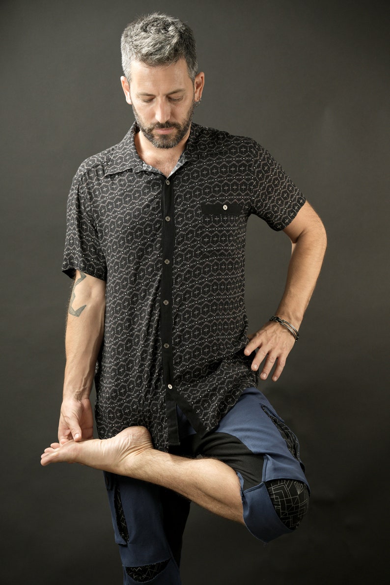 May include: A man wearing a black and white patterned short-sleeved shirt and blue trousers with a geometric design. He is standing with one leg bent and his foot resting on his thigh.