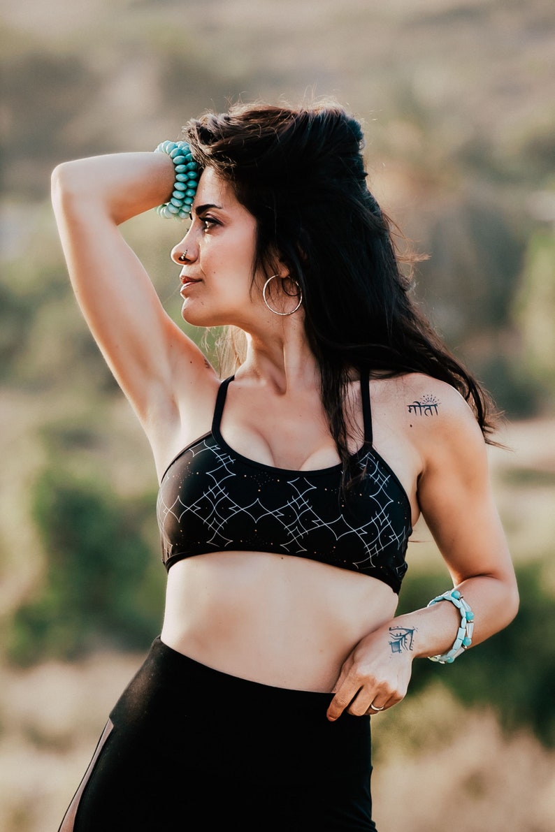 Peut inclure: Une femme portant un soutien-gorge de sport noir &agrave; motif g&eacute;om&eacute;trique et un legging noir. Elle a un petit tatouage sur l'&eacute;paule qui dit "माता" en hindi. Elle porte des bracelets turquoise aux deux poignets.