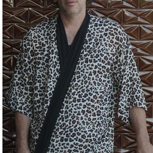 May include: A kimono-style robe with a leopard print pattern in brown and black spots on a cream background. The robe has a black lapel and is worn with a black shirt. The person is wearing a brown hat and sunglasses.