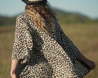 Pode incluir: Uma pessoa usando um chapéu preto com uma faixa estampada e um top estilo quimono creme. O top apresenta um padrão de estampa de leopardo marrom. O fundo é uma cena externa desfocada com tons verdes e marrons.