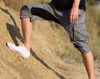 Op de afbeelding: Grijze cargoshorts met zwarte accenten en zakken. De shorts hebben een uniek ontwerp met een geometrisch patroon op de knieën. De persoon draagt witte sneakers en een zwart shirt. De shorts zijn knielang.