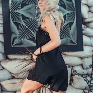 May include: A woman with long blonde hair wearing a black slip dress and black bracelets. She is standing in front of a wall with a black and white geometric design.