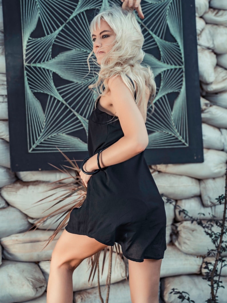 May include: A woman with long blonde hair wearing a black slip dress and black bracelets. She is standing in front of a wall with a black and white geometric design.