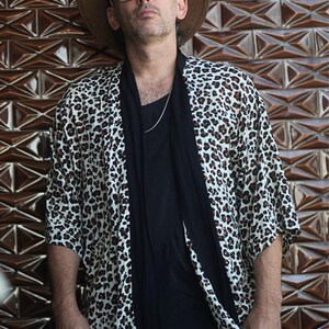 May include: A leopard print kimono-style jacket with a black shirt and a brown hat. The jacket has a white background with brown spots. The person is wearing sunglasses and a silver chain. The background is a brown, geometric tile wall.