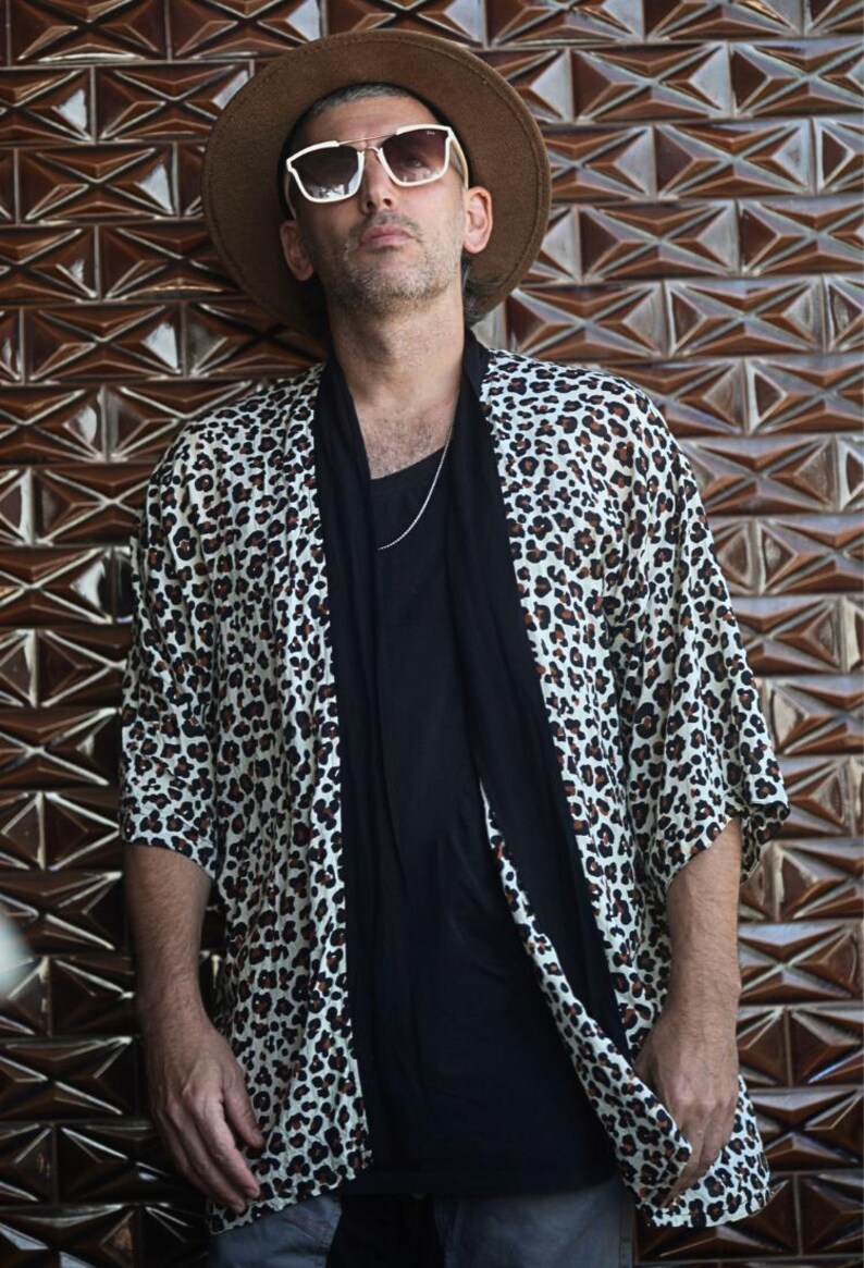 May include: A leopard print kimono-style jacket with a black shirt and a brown hat. The jacket has a white background with brown spots. The person is wearing sunglasses and a silver chain. The background is a brown, geometric tile wall.