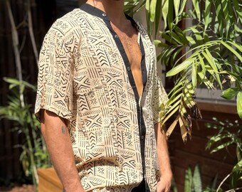 Op de afbeelding: Beige overhemd met een zwart geometrisch patroon en zwarte afwerking. Het shirt met korte mouwen heeft een opstaande kraag en is bij de borst opengeknoopt. Het shirt wordt gedragen met een zwarte broek.