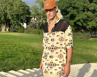Op de afbeelding: Een crème-kleurig overhemd met korte mouwen en een geometrisch patroon in zwart, bruin en bruin. Het shirt heeft een zwart mesh paneel op de schouders. De persoon draagt een bruine pet.