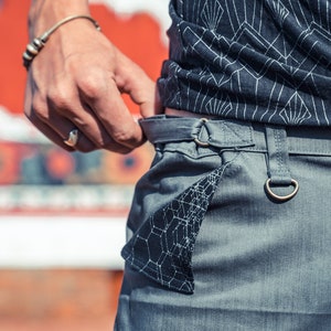 May include: Grey trousers featuring a herringbone texture, a black pocket flap with a white geometric web pattern, a metal D-ring attached to the waistband, and a black top with a white art deco-inspired line pattern.