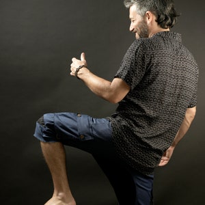 May include: A man wearing a black and white patterned short-sleeved shirt and blue denim shorts with a cargo pocket. He is standing with his right leg raised and his right hand raised in a thumbs up gesture.
