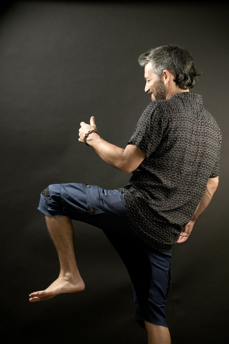 May include: A man wearing a black and white patterned short-sleeved shirt and blue denim shorts with a cargo pocket. He is standing with his right leg raised and his right hand raised in a thumbs up gesture.