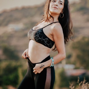 Peut inclure: Une femme portant un soutien-gorge de sport noir avec des motifs g&eacute;om&eacute;triques blancs et un legging noir avec un panneau transparent sur le c&ocirc;t&eacute;. Elle est debout &agrave; l'ext&eacute;rieur avec un arri&egrave;re-plan flou.