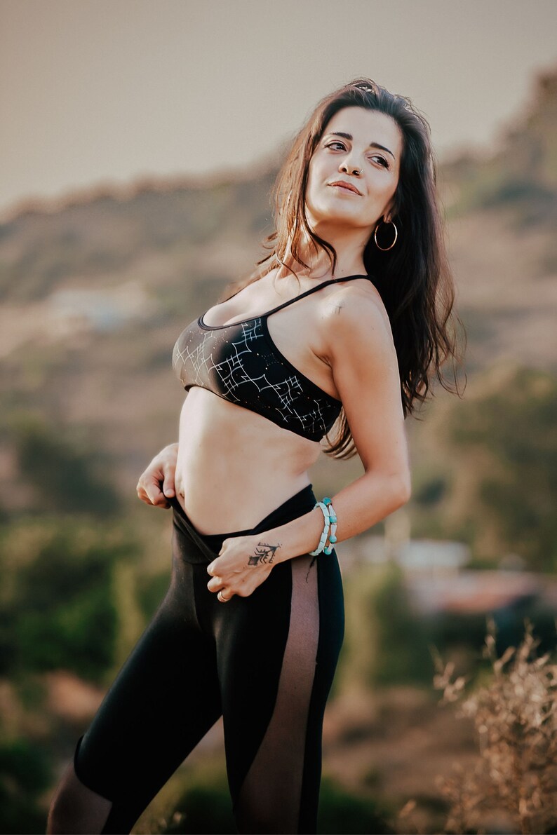 Peut inclure: Une femme portant un soutien-gorge de sport noir avec des motifs g&eacute;om&eacute;triques blancs et un legging noir avec un panneau transparent sur le c&ocirc;t&eacute;. Elle est debout &agrave; l'ext&eacute;rieur avec un arri&egrave;re-plan flou.