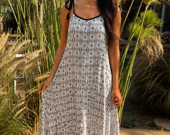 May include: A floor-length white dress featuring a repeating black geometric star pattern, thin black spaghetti straps with tie details at the shoulders, and a black trim along the neckline. The garment has a loose, flowing silhouette.