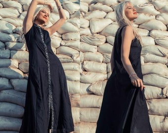 May include: A woman wearing a black sleeveless maxi dress with a silver zip detail. She is standing in front of a wall of white sandbags.
