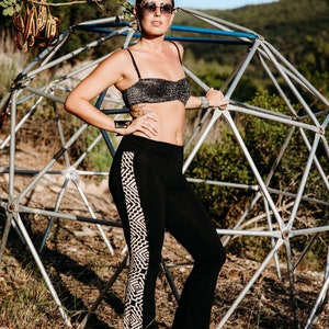 Puede incluir: Una mujer con pantalones negros con un patrón geométrico blanco en el lateral, un top bandeau negro brillante y gafas de sol. Está de pie frente a una estructura metálica.