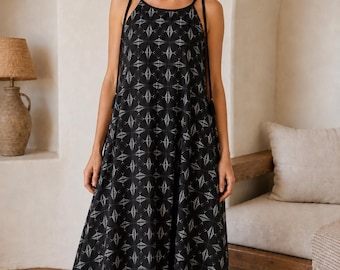 May include: A floor-length black sleeveless dress featuring a repeating white geometric diamond pattern. The garment has a high neckline with thin black shoulder straps that extend down the sides, ending in small black tassels at the waistline.