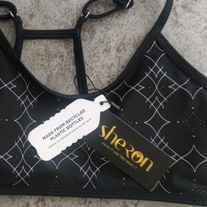 Peut inclure: Soutien-gorge de sport noir avec un motif g&eacute;om&eacute;trique blanc et des accents dor&eacute;s. Le soutien-gorge est fabriqu&eacute; &agrave; partir de bouteilles en plastique recycl&eacute;es. L'&eacute;tiquette indique "sheron wear it like you mean it".