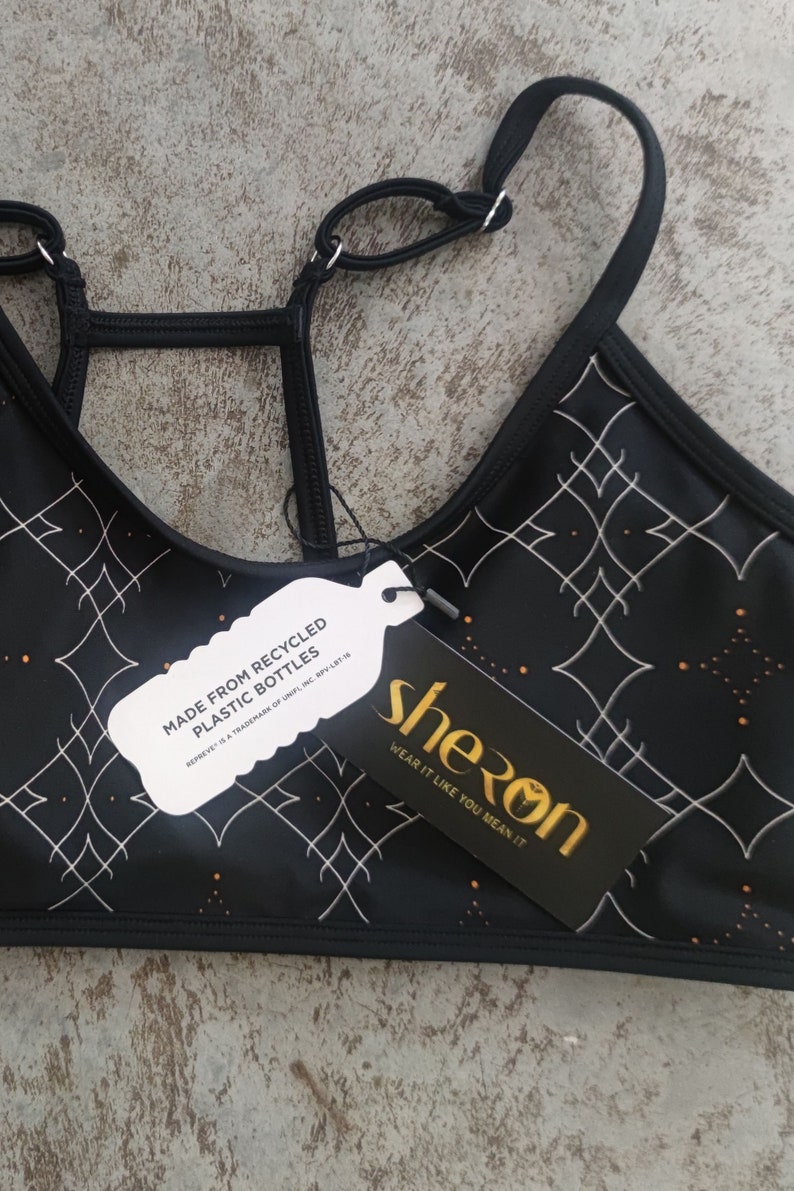Peut inclure: Soutien-gorge de sport noir avec un motif g&eacute;om&eacute;trique blanc et des accents dor&eacute;s. Le soutien-gorge est fabriqu&eacute; &agrave; partir de bouteilles en plastique recycl&eacute;es. L'&eacute;tiquette indique "sheron wear it like you mean it".