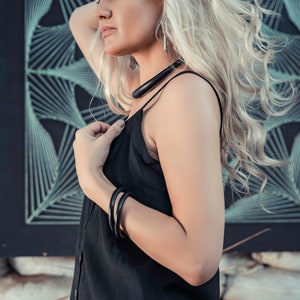 May include: A woman with long, blonde hair wearing a black sleeveless dress and a black necklace. She is standing in front of a black and white geometric pattern.
