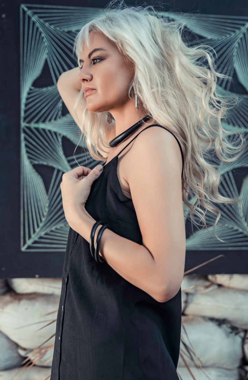 May include: A woman with long, blonde hair wearing a black sleeveless dress and a black necklace. She is standing in front of a black and white geometric pattern.