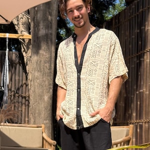 Może przedstawiać: Beżowa koszula zapinana na guziki z czarnym kołnierzem i wykończeniami, z geometrycznym wzorem w kolorze czarnym. Koszula z krótkim rękawem jest noszona z czarnymi spodniami. Osoba uśmiecha się, stojąc na zewnątrz.