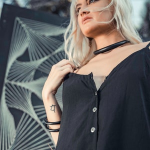 May include: A woman with blonde hair wearing a black sleeveless button-down top with a black choker necklace. She has a tattoo on her wrist.