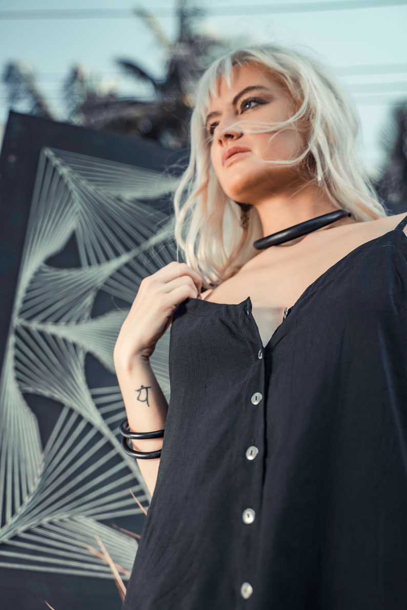May include: A woman with blonde hair wearing a black sleeveless button-down top with a black choker necklace. She has a tattoo on her wrist.