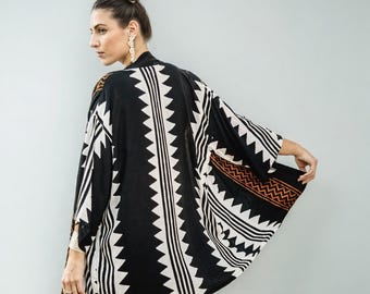 Op de afbeelding: Een zwarte kimono met een opvallend geometrisch patroon in wit en oranje. De kimono heeft een zigzagontwerp in het midden en langs de randen, met horizontale strepen en oranje accenten. De kimono is gemaakt van een zachte, vloeiende stof.