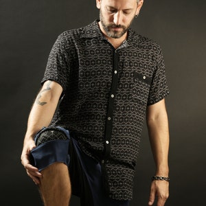 May include: A man wearing a black and white patterned short-sleeved shirt with a button-down collar and blue trousers. He is standing with his left leg bent and his right leg straight.