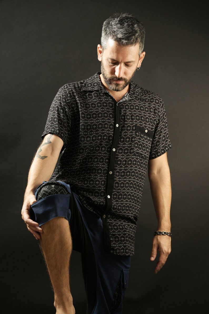 May include: A man wearing a black and white patterned short-sleeved shirt with a button-down collar and blue trousers. He is standing with his left leg bent and his right leg straight.