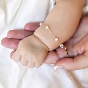 May include: A delicate gold chain bracelet featuring small white pearls spaced evenly along the length. The bracelet includes a small gold cross charm dangling from the end, creating a simple and refined piece of jewelry designed for a small wrist.