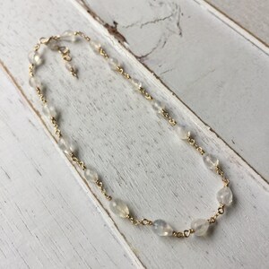 May include: A delicate gold chain necklace with small, clear moonstone beads. The necklace is on a white wooden surface.