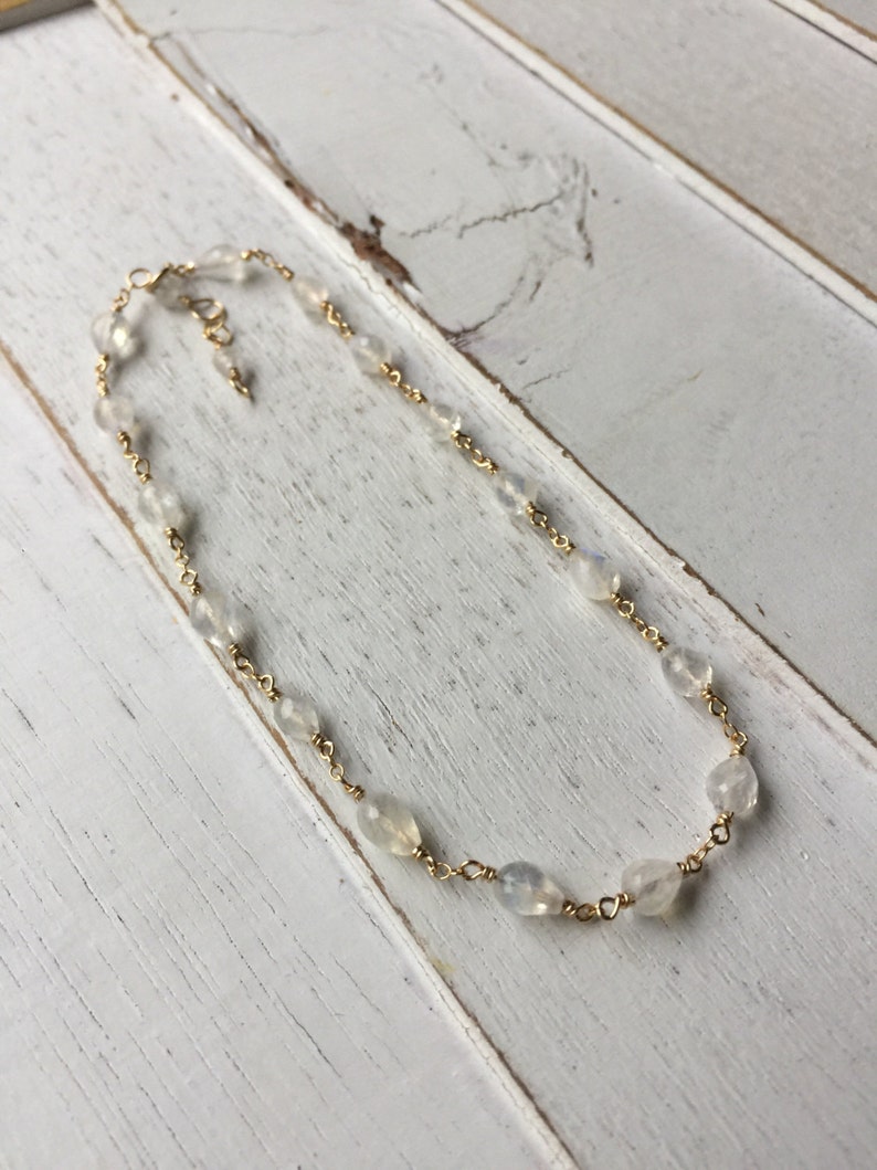 May include: A delicate gold chain necklace with small, clear moonstone beads. The necklace is on a white wooden surface.