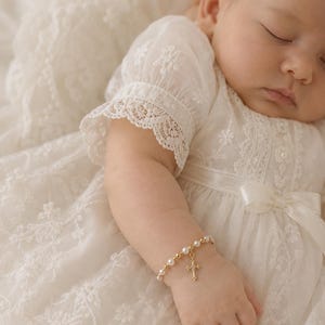 May include: A delicate gold-toned bracelet featuring small white beads and a central cross charm, worn on a wrist. The bracelet is paired with a cream-colored dress made of sheer fabric with intricate white floral embroidery and lace trim details.