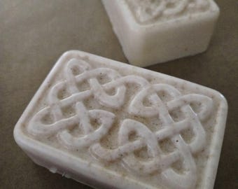 May include: Two rectangular bars of off-white soap with a speckled texture. Each bar features a raised, embossed Celtic knot design on the top surface, characterized by intricate, continuous interlacing lines forming a symmetrical pattern.
