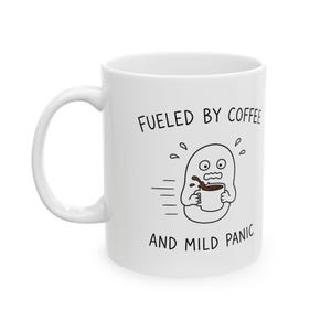 May include: A white ceramic mug featuring a black line drawing of a wide-eyed, sweating character holding a coffee cup with spilled liquid. The text above reads FUELED BY COFFEE and the text below reads AND MILD PANIC in black font.