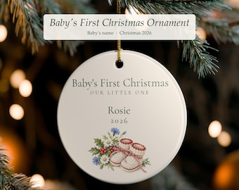 Può includere: Un ornamento rotondo in ceramica bianca appeso a un cordino dorato. Il testo recita Baby's First Christmas, OUR LITTLE ONE, Rosie e 2026. Sotto il testo c'è un'illustrazione colorata di fiori bianchi e blu, foglie verdi e scarpine da neonato.