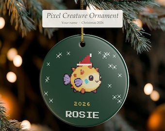 Può includere: Una decorazione natalizia rotonda verde scuro con un pesce palla in pixel art che indossa un cappello da Babbo Natale rosso. Il design include piccoli fiocchi di neve pixelati bianchi, l'anno 2026 e il nome ROSIE in testo pixelato bianco.