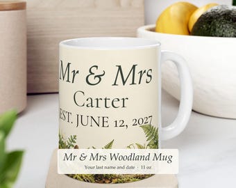 Può includere: Una tazza in ceramica bianca con il testo verde scuro Mr & Mrs Carter EST. JUNE 12, 2027. La base della tazza è decorata con un'illustrazione dettagliata di felci verdi, piccole pigne e fogliame boschivo su uno sfondo color crema.